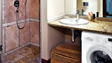 Wnętrza. Kamienny blat z umywalką służy również jako półka na kosmetyki. Inne rzeczy są trzymane w kuferku stojącym obok pralki. Wnętrze oświetla sufitowa lampa z trzema ruchomymi reflektorkami i kinkiety zamontowane na lustrze.