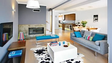 Dom Justyny i Krzysztofa miał być jasny i przestronny. Nowoczesny, ale w żadnym razie nie chłodny. Praktyczny, ale nieprzeładowany przedmiotami. a do tego utrzymany w delikatnych, uspokajających barwach, które łatwo urozmaicić kolorowymi dodatkami. <BR /> WNĘTRZA. Ściany w salonie pomalowano na dwa odcienie szarości. Betonowy fragment wokół kominka pokryto żywicą, stąd ciekawy wzór i kolor. Kąt wypoczynkowy oświetla lampa wisząca, przypominająca nieco przeskalowaną lampę biurkową, oraz zabawna lampka-łowiczanka, autorstwa polskiej projektantki Katarzyny Herman-Janiec.