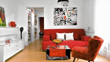 Z białymi ścianami kontrastują meble w mocnych kolorach - sofa (IKEA) i fotel z lat pięćdziesiątych w nowym obiciu (zdobycz z internetu). Abstrakcyjny obraz to po prostu kawałek czarno-białej tkaniny z IKEA, który gospodarze rozpięli na blejtramie i pokolorowali. Stolik kawowy jest dziełem projektantki; zrobiła go z płyty meblowej i 'ubrała' w pokrowiec i bieżnik z kieszeniami. Szufladom w białej komodzie dodano czerwone uchwyty z taśmy pasmanteryjnej - teraz łatwiej je otworzyć. aranżacja salonu, wystrój wnętrz