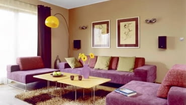Ściany w salonie mają ciepły kolor określany jako kawa z mlekiem. Od nich odcina się zestaw wypoczynkowy pokryty fioletową alcantarą i pomarańczowa lampa. Nad kanapą - grafiki 'Balet' autorstwa Joanny Burdy. UWAGA PONOWNA PUBLIKACJA ZA ZGODA WLASCICIELI