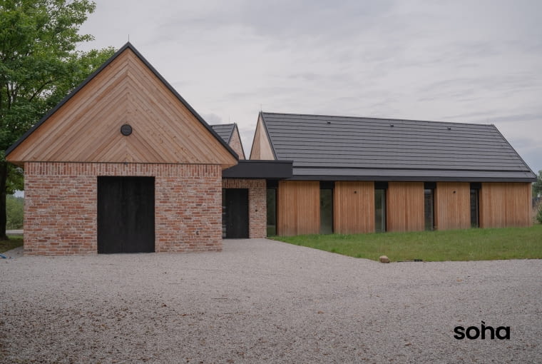 Murowano-drewniane elewacje przykrywa od góry ciemna, solidna linia dwuspadowych dachów, udanie kotwicząca lekkie bryły w konkretnej rzeczywistości. Tylko fragment centralny wyposażono w dach płaski Murowano-drewniane elewacje przykrywa od góry ciemna, solidna linia dwuspadowych dachów, udanie kotwicząca lekkie bryły w konkretnej rzeczywistości. Tylko fragment centralny wyposażono w dach płaski
