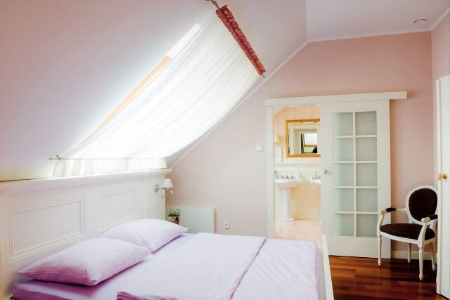Gospodarzom tej sypialni żaluzje lub zwijane rolety, stosowane na ogół do zasłaniania okien połaciowych, wydały się mało romantyczne. Poszukiwali bardziej stylowego rozwiązania, wybrali więc pomysłowo zamocowany zwiewny woal. <BR />DEKORACJE - NASTROJOWE OKNO NA PODDASZU. Aby zasłonka nie opadała na łóżko, kilkanaście centymetrów od dołu zrobiono tunel, w który wciągnięto drugi karnisz, zamocowany nieco nad parapetem. Bardzo pomysłowe, godne naśladowania rozwiązanie.