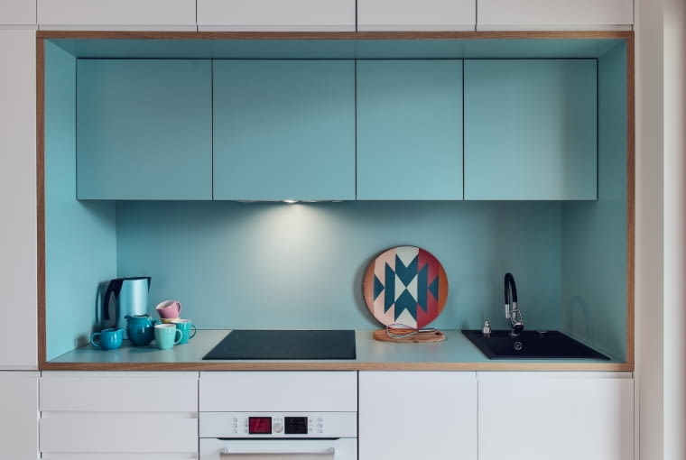 Otwarty na jadalnię aneks kuchenny ma świeże połączenie kolorystyczne bieli i turkusu. Wnękę pomalowano satynową farbą w tym samym odcieniu, co wiszące w niej szafki, tworząc jednolitą kompozycję.
