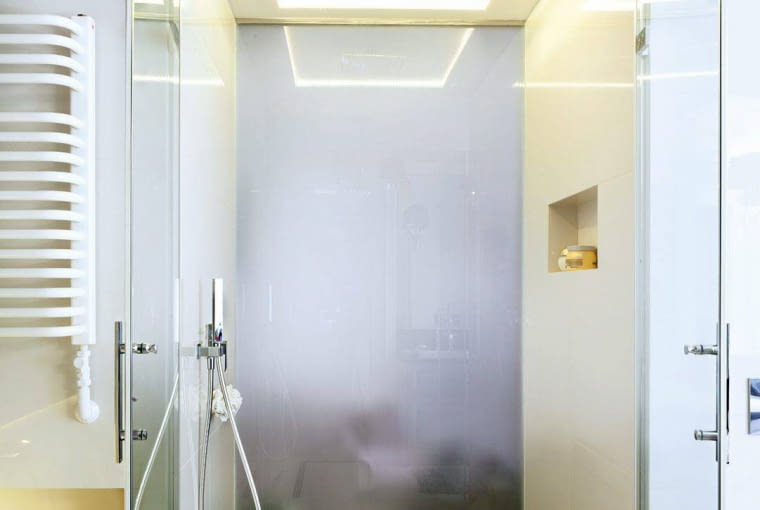 ŁAZIENKA. Czarna wstawka na podłodze kabiny to granit (z takiego samego zrobiono blat pod umywalkę). W wycięciu podwieszonego sufitu zainstalowano deszczownicę, a wokół niej - ledowe oświetlenie. Tylna ściana kabiny, sąsiadująca z sypialnią, jest zrobiona z matowego szkła.