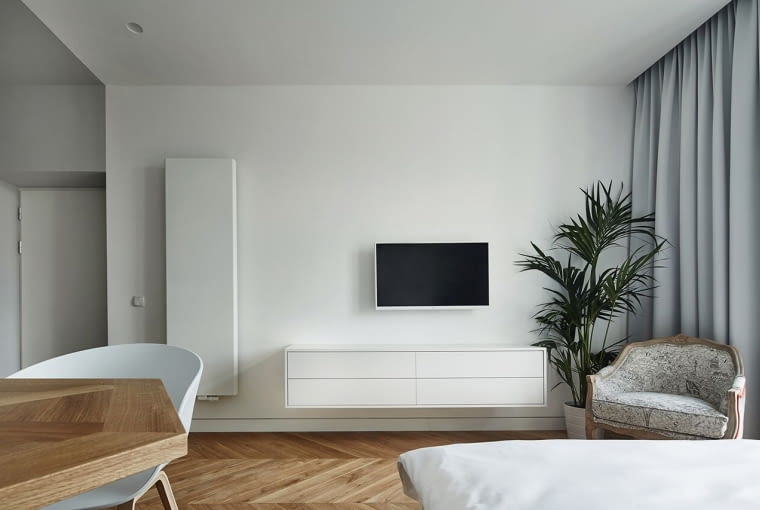 Jednolita przestrzeń służy również jako część dzienna, dlatego znalazł się w niej także kącik telewizyjny oraz wygody fotel. Wystrój apartamentu zmiękczają detale: szare, miękkie , lejące się zasłony oraz duży kwiat doniczkowy. małe mieszkanie, kawalerka, jak urządzić kawalerkę
