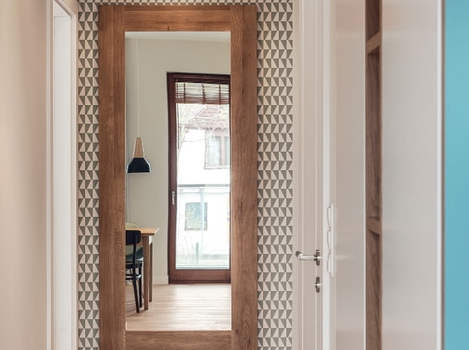 Obie części mieszkania łączy łamany korytarz. Duże lustro zawieszone naprzeciw kuchni i wyjścia na balkon odbija światło, optycznie powiększając powierzchnię. W tle na ścianie bestsellerowa tapeta firmy Boras 'Trapez' w trójkąty (trapezy). Geometryczny wzór stanowi niebanalną dekorację przedpokoju. 