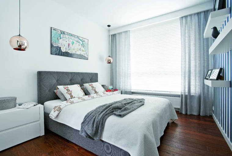 W SYPIALNI króluje szare łóżko z tapicerowanym zagłówkiem (Nap). Nad szafkami nocnymi wiszą lampy z miedzianymi kloszami (Ap-Design), zastępujące tradycyjne lampki nocne. Ściana oklejona tapetą w biało-niebieskie pasy przypomina gospodarzom o miłych chwilach spędzonych pod żaglami.