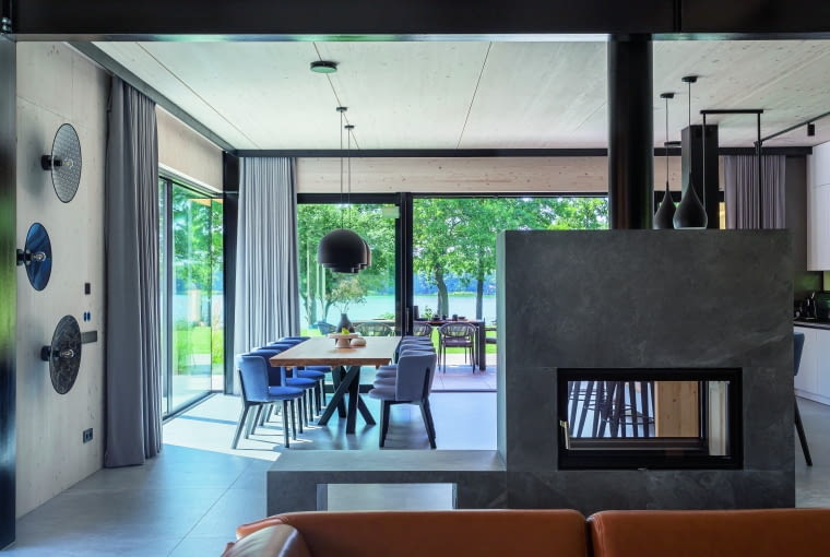 Widok z salonu na stół jadalniany oraz kominek na drewno, który dzieli otwartą strefę dzienną mniej więcej na połowę, a jest wykorzystywany okazjonalnie jako dodatkowe źródło ciepła. Po lewej trzy oryginalne lampy-kinkiety, przypominające artystyczną instalację Widok z salonu na stół jadalniany oraz kominek na drewno, który dzieli otwartą strefę dzienną mniej więcej na połowę, a jest wykorzystywany okazjonalnie jako dodatkowe źródło ciepła. Po lewej trzy oryginalne lampy-kinkiety, przypominające artystyczną instalację