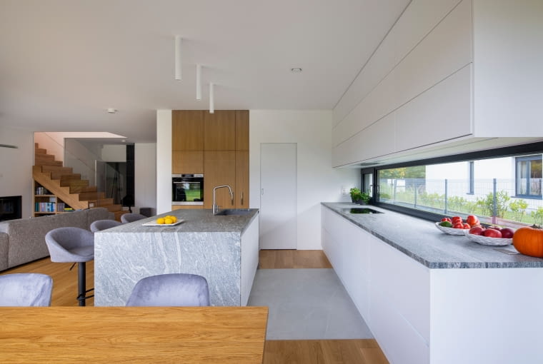 Wyspowe szarości szczotkowanego i płomieniowanego granitu Kuppam Green znajdują z kolei odbicie w sąsiednich kamiennych blatach roboczych usytuowanych wzdłuż wąskich, horyzontalnych okien kuchni – tu naprawdę jest gdzie się rozpędzić z przygotowywaniem potraw! Dom z kostki