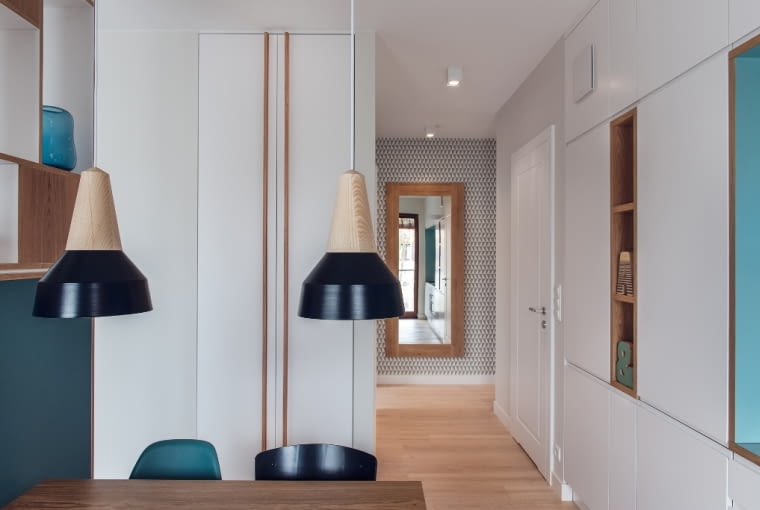 Z korytarza wchodzi się do otwartej przestrzeni części dziennej. Po środku znajduje się kącik jadalny, który czasem zamienia się w miejsce do pracy. Wykonany na wymiar regał przy stole to 'ścianka', oddzielająca jadalnię od małej sypialni właścicieli. Z prawej zlokalizowano aneks kuchenny.