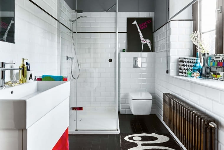 ŁAZIENKOWA KLASYKA, czyli białe płytki-cegiełki. Czarny pasek między nimi subtelnie nawiązuje do koloru podłogi. Maria jest dumna z tego, że udało jej się uratować oryginalny grzejnik i dobrać do niego kinkiet w tym samym odcieniu. Na podłodze dywaniki ze Scandinavian Living. Żółty dywanik przełamuje dwubarwną kolorystykę i ożywia nieco ascetyczny wystrój łazienki.