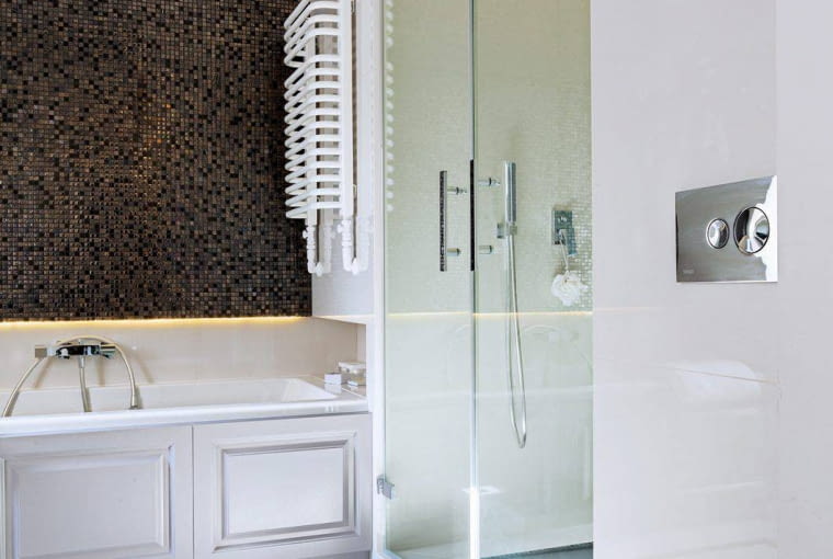 ŁAZIENKA. Największą dekoracją wnętrza jest kamienna mozaika, którą wyłożono ścianę nad wanną. Szare, czarne i perłowe kostki pięknie się mienią w świetle, zarówno naturalnym, jak i sztucznym. U dołu, w podcięciu ściany, zainstalowano ledowe oświetlenie. Wnęka pod grzejnikiem służy jako półka na kosmetyki.