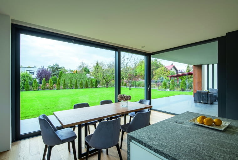Dzięki umieszczeniu jadalni w sąsiedztwie tarasowych podcieni wydaje się, jakby była ona osadzona w dodatkowym, przeszklonym kubiku – grafitowa kolorystyka profili okiennych świetnie łączy się tu z szarością welurowych obić krzeseł, przechodzącą w granitowe blaty wyspy Dom z kostki