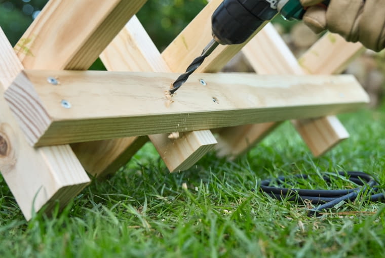 Krok 6. Zamocuj listwy zaciskowe i drugą deskę Rozłóż zawias rozwórki tak, aby elementy nóżek znajdowały się pod kątem 45°. Po ustawieniu koziołka do piłowania drewna przykręć listwy zaciskowe, każdą dwiema śrubami 4,5 × 70 mm – jedna powinna się znaleźć w dolnej części koziołka drewnianego, a druga – w górnej. Listwy zaciskowe ograniczają swobodę ruchu trzech drewnianych rozpórek. Na koniec zamocuj drugą, dolną deskę o długości 75 cm.