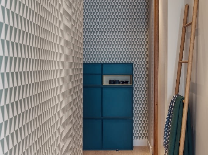 Obie części mieszkania łączy łamany korytarz. Duże lustro zawieszone naprzeciw kuchni i wyjścia na balkon odbija światło, optycznie powiększając powierzchnię. W tle na ścianie bestsellerowa tapeta firmy Boras 'Trapez' w trójkąty (trapezy). Geometryczny wzór stanowi niebanalną dekorację przedpokoju. 