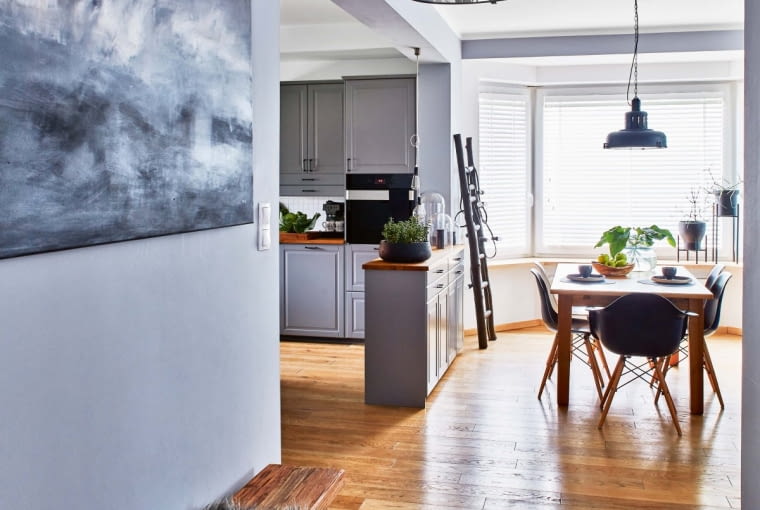 Płytkie szafki kuchenne pełniące funkcję barku symbolicznie odgradzają kuchnię od salonu. Na podłodze położono bielone dębowe deski. Na pierwszym planie wisi lampa z tej samej serii co ta nad stołem. Autorką obrazu jest Margo Hupert.