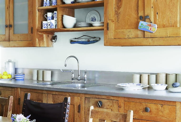 Kuchnia z szafkami o drewnianych frontach. corner of dining table, showing a plates and a bowl, stones and flowers, in the kitchen, side unit work surface with a sink, wooden units and cupboards, storage shelves with plates, cups, jars on the side. Picture by Country Living March 2008 / Retna UK Ref: CLV Local Caption 140 Pictures owned by Natmags Exceptions:Unknown Embargo:UK: No sales to Countryside, Coastal, and Homes Garden titles in UK. Minimum page rate of 250- for use 3 months after cover date Foreign (exc US): Yes for use 3 months after cover date US: No sales to house and gardens/interiors/countryside magazines Approval required for third party purchasers for usage on greeting cards, diaries, calendars.
