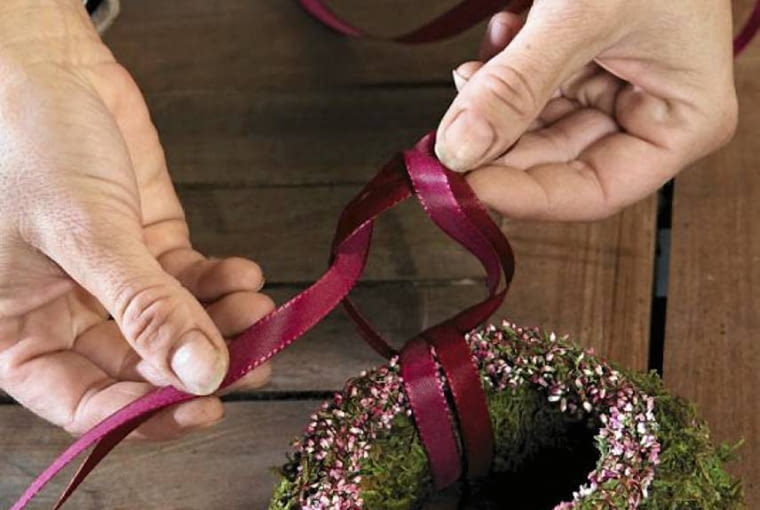 Wianuszek z wrzosu kładziemy na nieco większy wianek z mchu, związujemy wstążkami i przyczepiamy je do kompozycji. Wianuszek z wrzosu kładziemy na nieco większy wianek z mchu, związujemy wstążkami i przyczepiamy je do kompozycji.