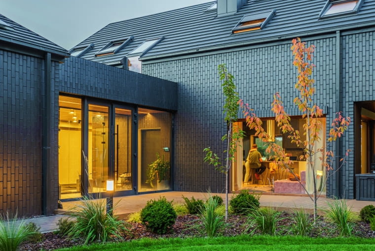 Dom stoi na planie litery L, ale gdy przyjrzeć mu się uważnie, widać, że w rzeczywistości tworzą go trzy bryły – mieszkalna (z prawej) oraz garażowa (z lewej), które zgrabnie łączy w całość parterowy moduł wejścia głównego, przekryty dachem płaskim Dom stoi na planie litery L, ale gdy przyjrzeć mu się uważnie, widać, że w rzeczywistości tworzą go trzy bryły – mieszkalna (z prawej) oraz garażowa (z lewej), które zgrabnie łączy w całość parterowy moduł wejścia głównego, przekryty dachem płaskim