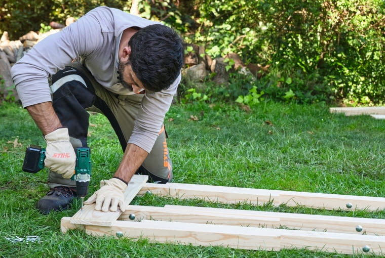 Krok 5. Włóż i przykręć pierwszą deskę Połóż deskę o długości 70 cm na już połączonych drewnianych listwach i przykręć ją do każdej listwy dwiema śrubami 4,5 × 70 mm. Upewnij się, że deska jest wyrównana ze środkową nogą.