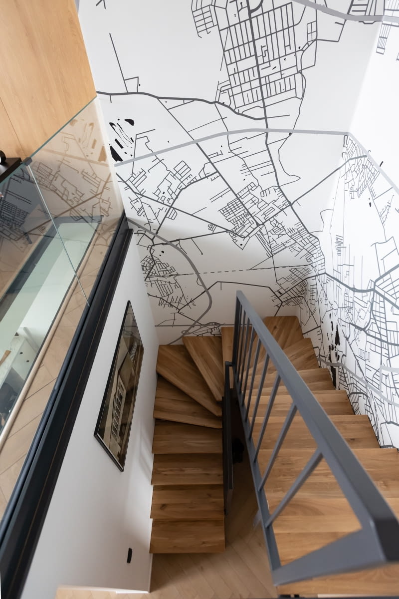 Tapeta z mapą Poznania, przyklejona do ścian w strefie wejścia i schodów na piętro, idealnie obrazuje zainteresowania i fascynacje właścicieli. Prezentuje motyw przewodni mieszkania, jakim jest ich rodzinny Poznań. Tapeta marki Ubierz Swoje Ściany przedstawia czarno-białą siatę ulic miasta, na której dostrzec można również punkt z lokalizacją prezentowanego mieszkania. Jak zauważa pani domu, przyjemnym zajęciem, zwłaszcza z poziomu sypialni, jest poszukiwanie charakterystycznych miejsc na mapie, co często robią dzieci lub goście.
