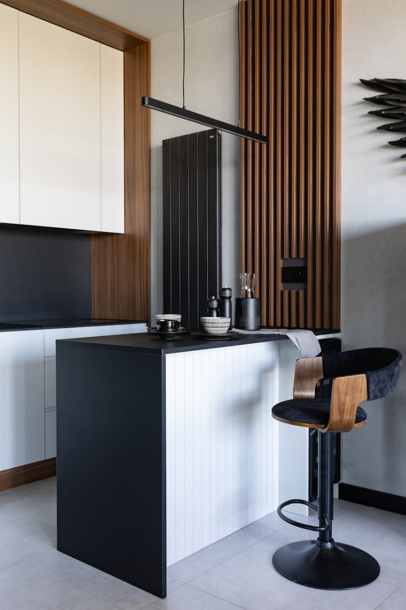Otwarta na salon kuchnia naśladuje styl i kolorystykę aranżacji całej strefy dziennej. Cechują ją proste, jak w całym wnętrzu, formy oraz wspólna paleta barw: szarość, czerń, biel i drewno. Nawet ściana nad blatem jest pozbawiona cech typowych dla kuchennych aranżacji. Zamiast tradycyjnych kafli wykończono ją tym samym, czarnym materiałem co blat kompaktowy. Ścianę przy wyspie kuchennej zdobi panel z lamelami pokrytymi laminatem o wzorze drewna. Formę pionowych listew powtarza front wyspy widoczny od strony salonu oraz grzejnik.