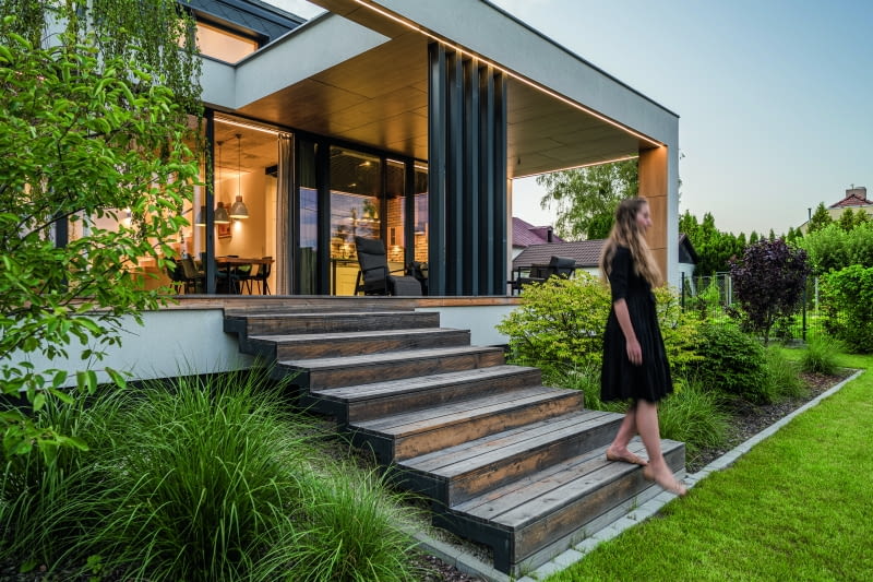 Z tego tarasu, na dolnej kondygnacji, można już schodzić do ogrodu bezpiecznie, nawet z lekko przymkniętymi oczami czy na bosaka  – zwłaszcza że dużą część przestrzeni przed domem zajmuje wypielęgnowany trawnik, którego systematycznego zraszania strzeże automatyka