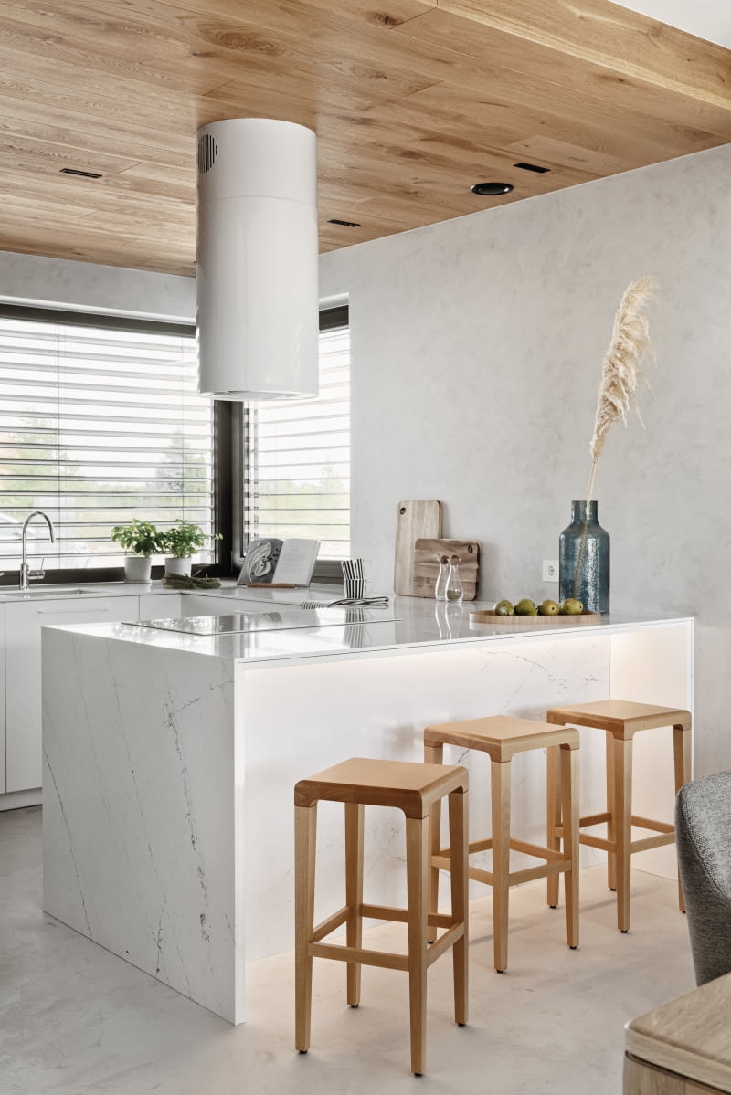 W kuchni dla uzyskania minimalistycznego efektu zrezygnowano z tradycyjnych płytek. Ściany wykończono więc tak samo jak podłogę – żywicą. Szafki powstały z białych, lakierowanych płyt oraz wspomnianych grafitowych płyt marki Forner. Ciemne fronty są wykonane w technologii anti-fingerprint, zapobiegającej powstawaniu na ich powierzchni odcisków palców. Na blaty użyto konglomeratu. A sufit zdobią dębowe deski, użyte w kilku innych miejscach domu.