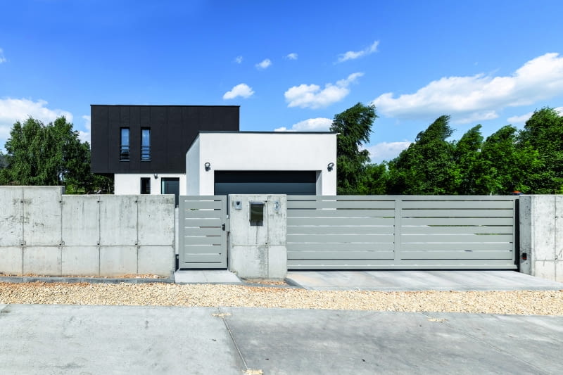 Kolorystyka domu to wyrazisty kontrast bieli parteru i czerni piętra, ale jeszcze większe  zróżnicowanie wprowadza ogrodzenie, wykonane z mocno surowego betonu, które zdecydowanie odcina się od oszczędnej elegancji nowoczesnej architektury jednorodzinnej