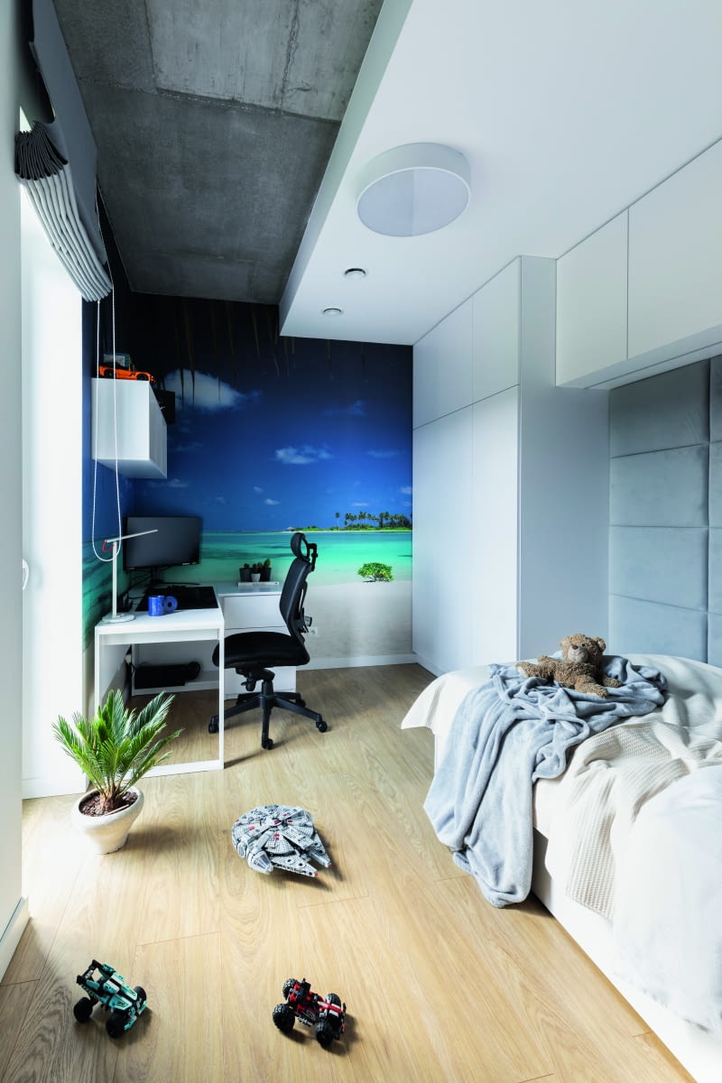 Drugi pokój dziecięcy to wyraźnie „chłopięce klimaty”, w dodatku w wydaniu dla nieco starszych. Widać podobny pomysł na pluszowe zagłówki  przy łóżku, jest też duży ścienny element graficzny, ale zabawki i wyposażenie bardziej grawitują już w kierunku techniki, włącznie z tą kosmiczną. Sufit bezceremonialnie wykończono zaś zupełnie dorosłym motywem. A jakże – betonowym