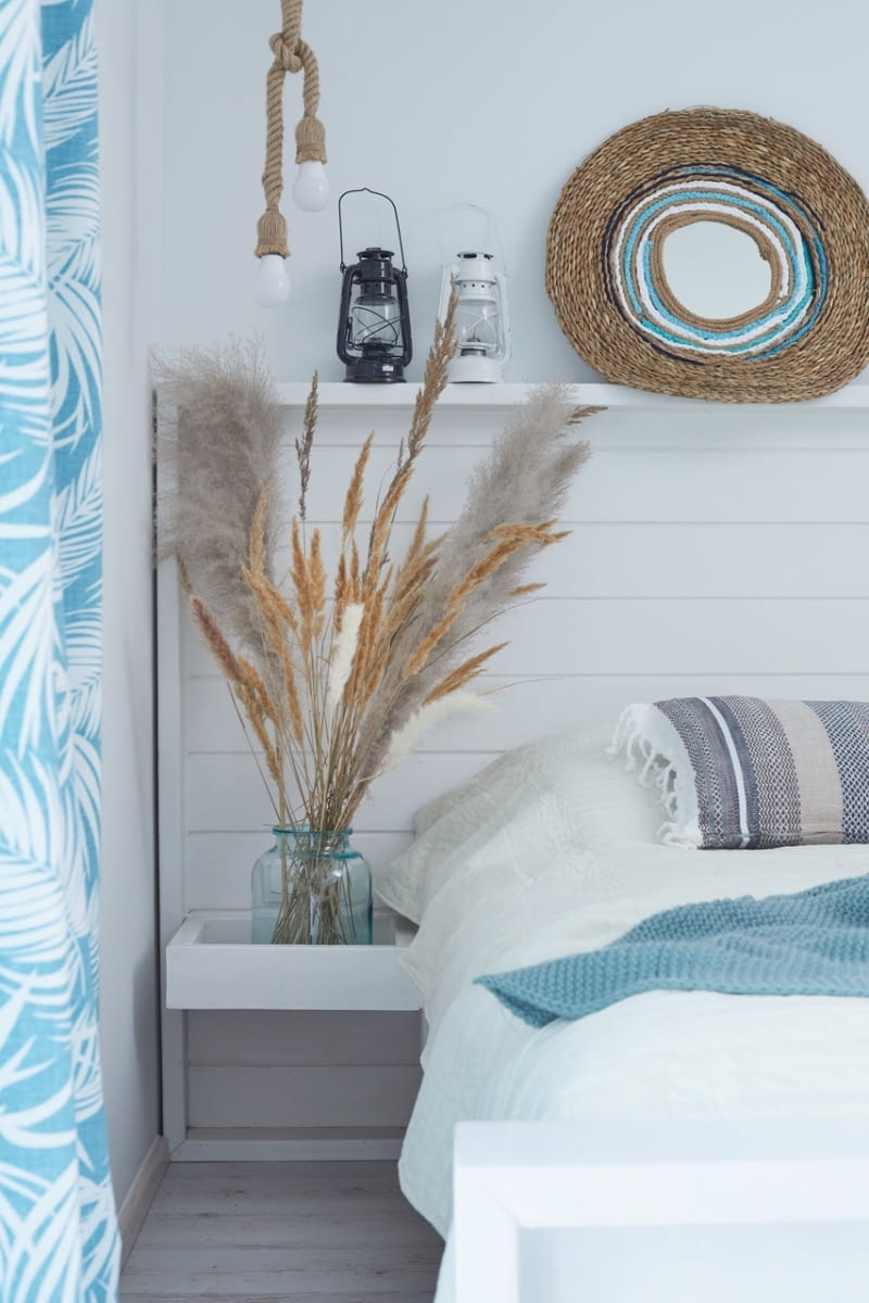 Wieczorne czytanie książek ułatwiają dwie wiszące po bokach materaca lampy z jutowych lin. Lusterko w oplocie z konopnych sznurków, lampy-latarenki i trawy dopełniają swobodny, niemal wakacyjny wystrój.