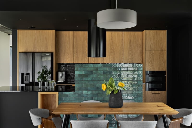 Czerń ścian i sufitu w strefie kuchni stanowi eleganckie tło dla wyrazistego rysunku forniru na frontach mebli. Paletę kolorystyczną subtelnie uzupełnia zieleń płytek, ułożonych na murku oddzielającym kuchnię od jadalni. Czerń ścian i sufitu w strefie kuchni stanowi eleganckie tło dla wyrazistego rysunku forniru na frontach mebli. Paletę kolorystyczną subtelnie uzupełnia zieleń płytek, ułożonych na murku oddzielającym kuchnię od jadalni.