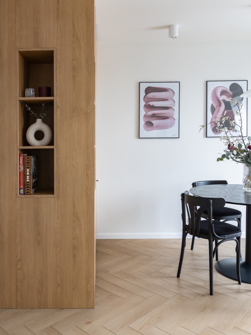 W niewielkiej przestrzeni jadalni idealnie sprawdza się okrągły stół ze sklepu sfmeble.pl. Towarzyszą mu krzesła Thonet po renowacji wykonanej przez Retro Wnętrza. Czarne meble przełamują górującą we wnętrzu biel i pięknie komponują się z drewnem kubika. Ozdobą zakątka są obrazy Nikodema Szpunara zakupione w sklepie sztuki.waw.pl. Zawierają nutę bordowej barwy – kolorystycznego motywu aranżacji całego mieszkania.