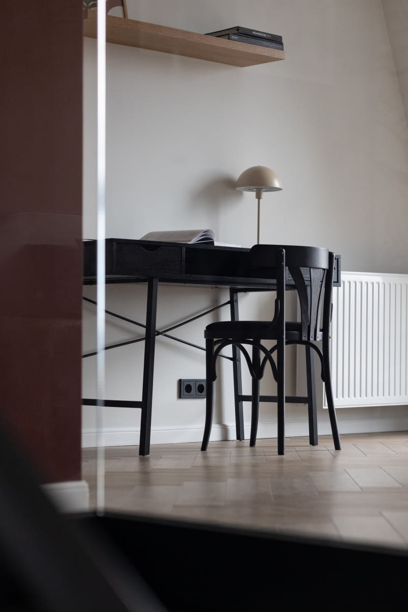 W zaciszu sypialni ustawiono też biurko do pracy. Wybrano model mebla o lekkiej konstrukcji, który nie ingeruje zanadto w przestrzeń pomieszczenia. Czarne, drewniane biurko Gavle wraz z odrestaurowanym krzesłem Thonet nawiązują do jadalnianej scenerii. W całym mieszkaniu zastosowano panele winylowe w odcieniu przygaszonego dębu Sava Herringbone Sand, ułożone w klasyczną jodełkę. Wspólna na parterze i piętrze, jasna podłoga pozwala uzyskać spójny wystrój, ładnie korespondujący z jasną paletą barw dobraną do aranżacji.