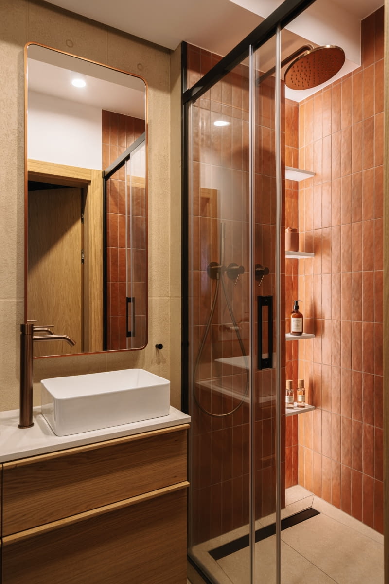 W łazience z prysznicem prym wiodą ciepłe tonacje rudości i drewna. Wnętrze kabiny prysznicowej wyłożono niedużymi płytkami Argile Cotto 6 x 24,6 cm producenta Equipe. Ich nazwa nie jest przypadkowa, bo z języka francuskiego oznacza „glinę”, co obrazuje, skąd cała kolekcja czerpie inspirację. Do przyjemnej barwy kafli dopasowano baterie Tremme  oraz ramę lustra w miedzianym odcieniu.