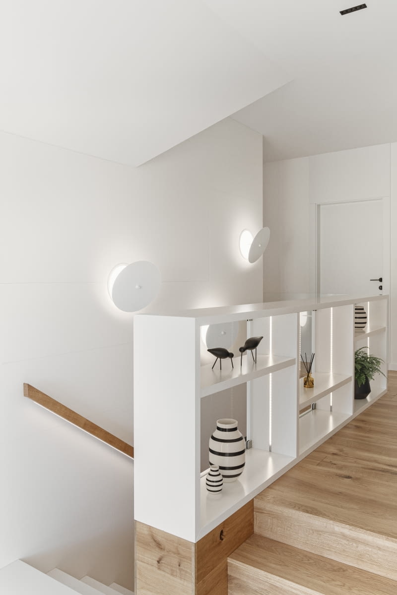 Pierwszy bieg schodów prowadzących na piętro wykończono białym konglomeratem kwarcowym o grubości 2 cm. Na drugim, po pokonaniu spocznika między piętrami, nawierzchnia zmienia się na drewnianą deskę dębową, obecną w korytarzu i pokojach domowników. Barierka schodów przyjmuje atrakcyjną formę białych, ażurowych półek, na których można trzymać dekoracyjne dodatki lub książki. Od strony schodów jest zabezpieczona hartowanym szkłem.