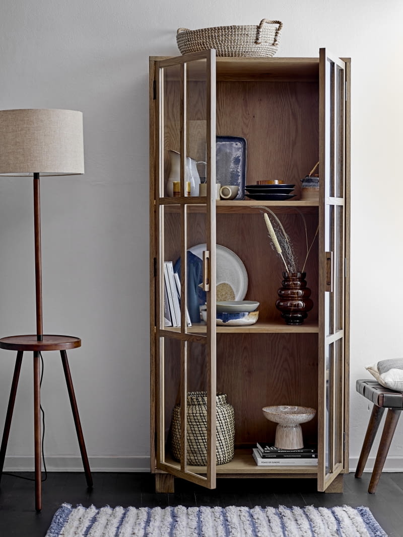 We wnętrzu przeszklonej, wysokiej szafki, ustawione obok siebie przedmioty o różnych wielkościach, kształtach i materiałach tworzą małe, dynamiczne komplety. Regulowane półki pozwalają zmieścić także większe elementy, a te najcięższe najlepiej jest umieścić na dole, by zachować stabilność i porządek kompozycji.