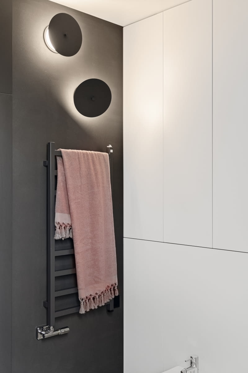 Kompozycja kolorystyczna w całym domu opiera się na kontrastach, dlatego również w łazienkach użyto tych samych środków wyrazu. Ściany i sufit w łazience dzieci mają grafitowy kolor. Pasujący do nich grzejnik i kinkiety również dobrano w tym odcieniu szarości. Ciemne ściany pokrywa żywica, a jej jasna wersja znalazła się na obudowie wanny i niskiej ściance kolankowej. Obudowę stelaża toalety i bidetu wraz z zaprojektowanymi nad nimi półkami stolarz przygotował z białych, lakierowanych płyt.