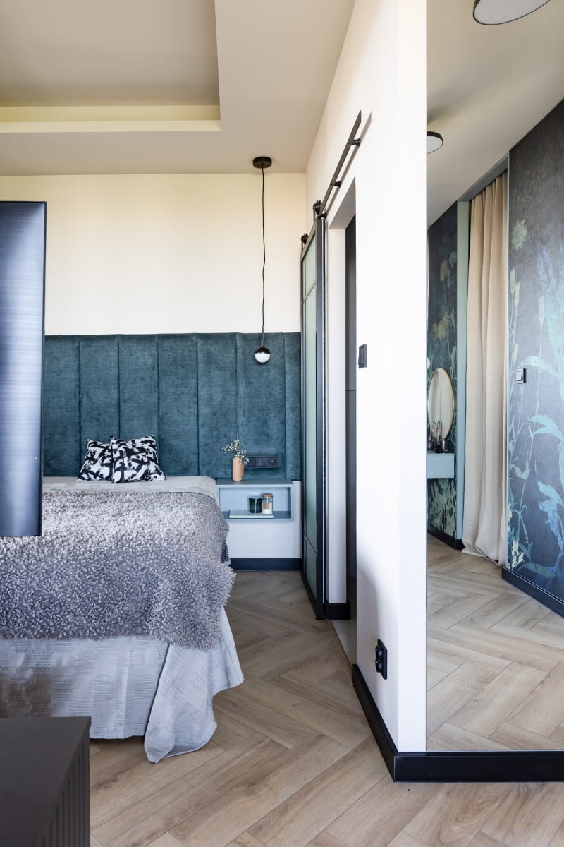 Tuż przy wejściu do sypialni na ścianie zamontowano lustro. Sprawdza się w tym miejscu zwłaszcza ze względu na bliskość garderoby. Pozwala przejrzeć się od stóp do głów, z możliwością wygodnego oddalenia się na parę kroków od tafli. Warto zwrócić też uwagę na podłogę pomieszczenia. Szare płytki ze strefy dziennej przechodzą tu w panele laminowane firmy Weninger układane w jodełkę. Deski o wyglądzie tradycyjnego parkietu dodają wnętrzu znajomego ciepła, które gwarantuje tylko drewno.