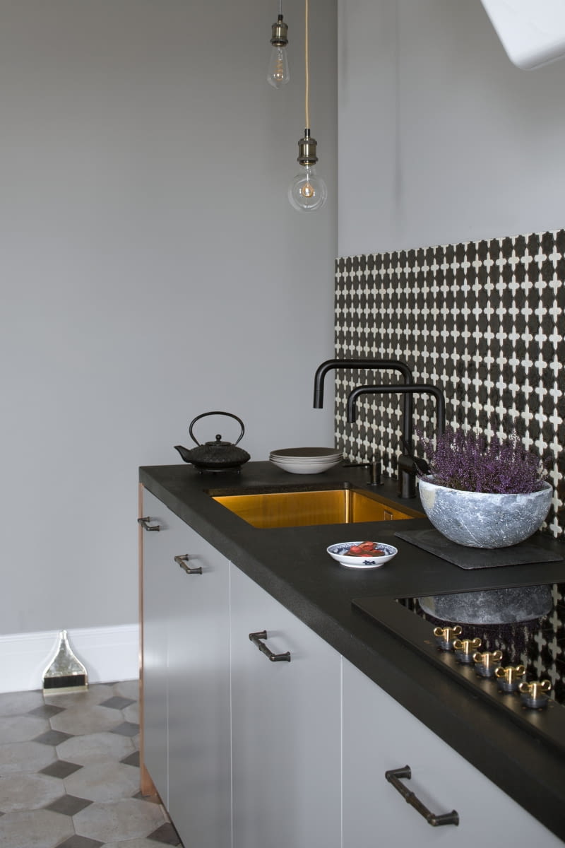 Na tle stonowanych, matowych, szarych frontów zabudowy kuchennej i ścian w tym kolorze mocno wybrzmiewają czarne, granitowe blaty i ułożona nad nimi mozaika z manufaktury Ardea oraz miedziana konstrukcja opleciona kablami z żarówkami. Miedziany jest także zlewozmywak Nivito. Na tle stonowanych, matowych, szarych frontów zabudowy kuchennej i ścian w tym kolorze mocno wybrzmiewają czarne, granitowe blaty i ułożona nad nimi mozaika z manufaktury Ardea oraz miedziana konstrukcja opleciona kablami z żarówkami. Miedziany jest także zlewozmywak Nivito.