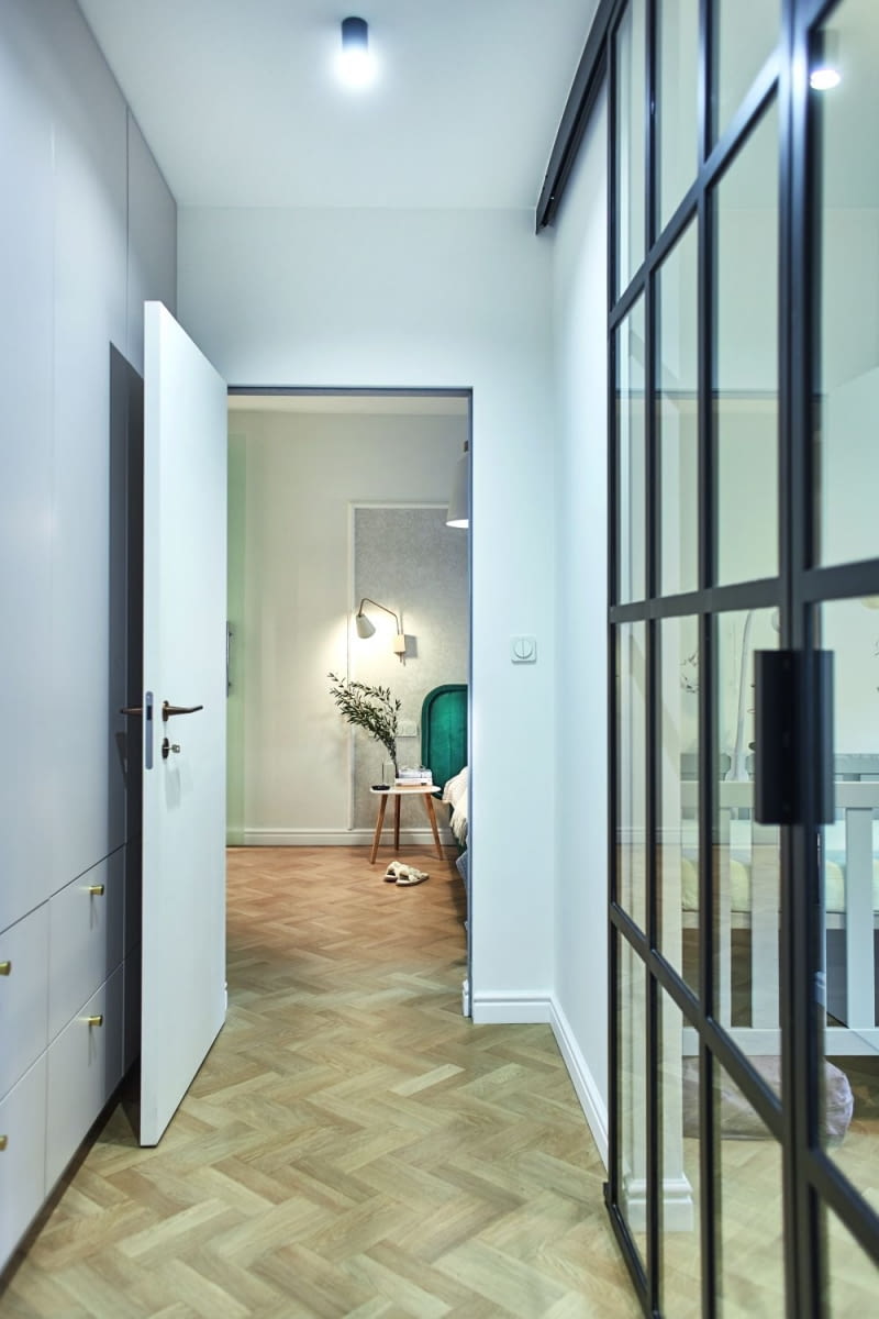 Korytarz prowadzący do sypialni rodziców. Loftowa ścianka dostarcza naturalne światło do holu, dzięki czemu nie jest on zupełnie ciemnym zakamarkiem, ale dość jasnym korytarzem, którym miło jest przechodzić do kolejnych pomieszczeń. Po lewej stronie, przy ścianie, zmieściła się wysoka do sufitu szafa. Jej fronty wykonano z tego samego co inne, jasne meble do zabudowy w mieszkaniu, z laminatu Kaszmir marki Egger. Ma odcień zbliżony do szarości.