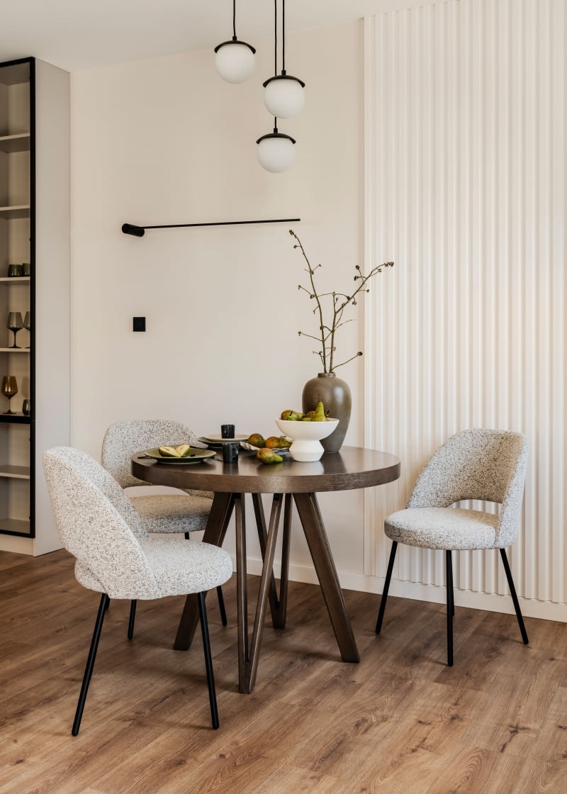 Tłem jadalnianej kompozycji jest nie tylko okno ujęte w kremową roletę i beżowe zasłony (dekoracje okienne Joanna Fibich zrealizowała we współpracy z Martą Pawlak z MP Interiors), ale też ściana wykończona białymi panelami z Mardom Decor, które tworzą subtelną grę światłocieni. Rozkładany stół z fornirowanym blatem to mebel producenta Bomar Meble, krzesła marki Halex Meble, a nad całością zawieszono lampę od Ummo Lighting. Kompozycję dopełnia czarny, minimalistyczny kinkiet od Nowodvorskiego.