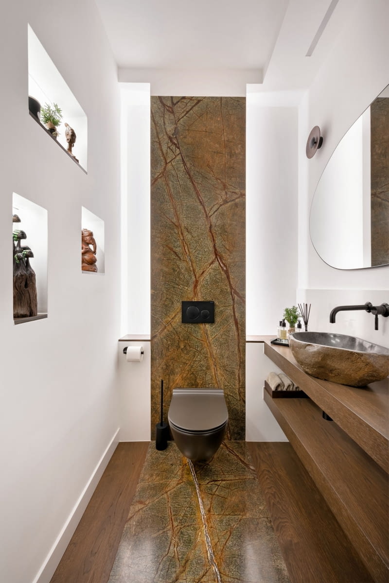 W toalecie, choć to często traktowane po macoszemu pomieszczenie, nie zabrakło oryginalnych rozwiązań. Zachwyca tu widoczny na ścianie i podłodze marmur Forest Green ze sklepu Prodmar. Po bokach kamienia na podłodze ułożono deski. Wykonany z zabezpieczonego przed wilgocią forniru blat dźwiga kamienną, 30-kilogramową umywalkę, zrobioną z rzecznego otoczaka. We wnękach ściennych stanęły figurki przywiezione z Kenii, Indii i Tajlandii.