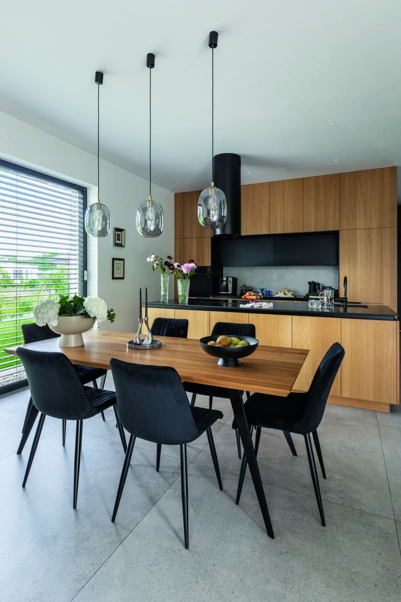 Jadalnia z widokiem na kuchnię – dobrze widać tu przemyślane i stylowo  prezentujące się zestawienia jasnego dębu zabudowy i stołu z czarnymi  aplikacjami krzeseł oraz granitu wyspowego blatu czy pochłaniacza  zapachów. Do tego te fantazyjne bańki wiszących lamp Kaspa ONYX!