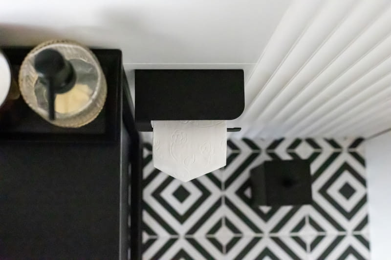 Czarno-biały wzór gresu Paradyż Modern motyw B na posadzce odnosi się do tapety przy schodach i stanowi główny element ozdobny pomieszczenia. W kontraście do podłogowych kafli, ściany łazienki pokryto białymi, matowymi płytkami Grespania Stark Blanco. Czarne akcesoria łazienkowe: uchwyt na papier toaletowy, szczotka WC i półka pod prysznicem – polski producent Dabstory