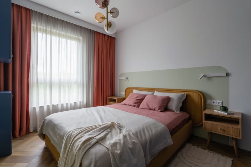 W sypialni atmosferę definiują miękkie tekstylia – to one nadają przestrzeni spokojny, otulający klimat. Zestaw pościeli z naturalnych włókien i lekkie narzuty wprowadzają subtelny ton ciepła. Najbardziej wyrazistym elementem są jednak okna ubrane w dwa rodzaje tkanin: masywniejsze zasłony w rdzawym tonie oraz delikatną, półprzezroczystą firanę. Przyjemne w dotyku, tapicerowane łóżko od Scandicsofa zaprasza do odpoczynku.