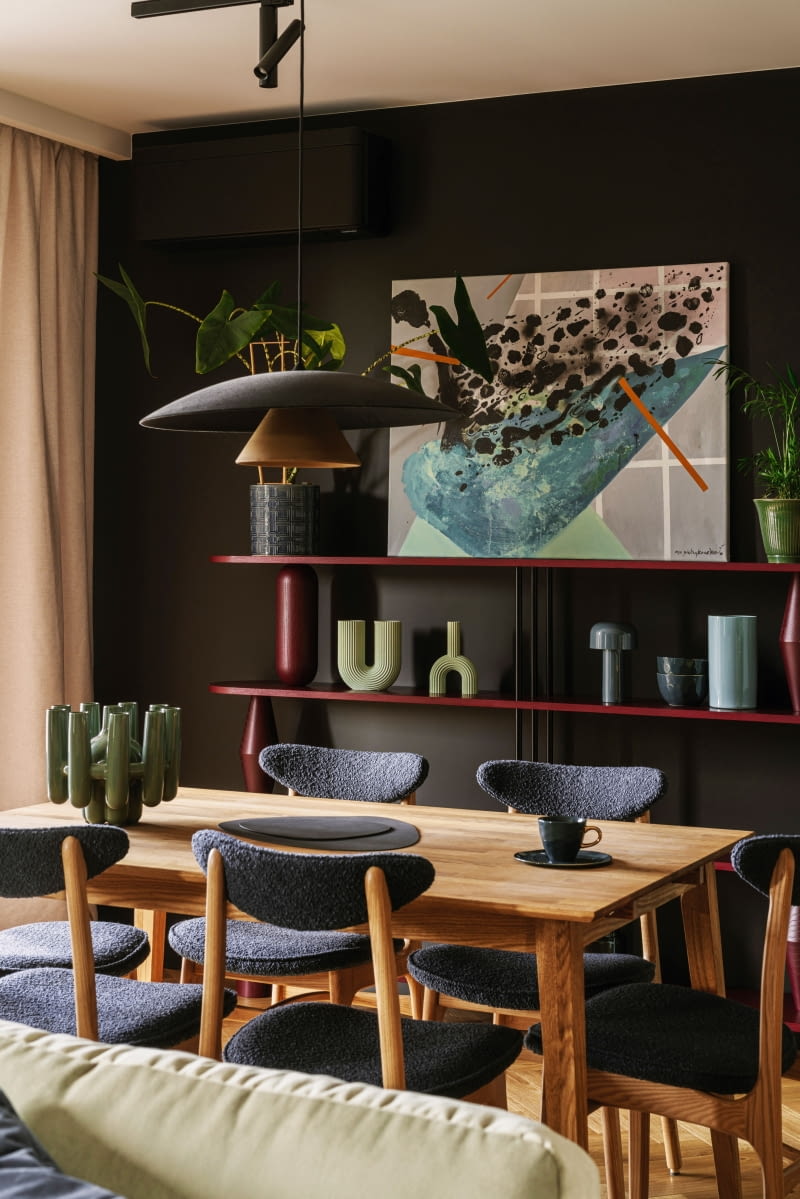 Bordowy regał marki Tamo stanowi jedno z najważniejszych, przyciągających uwagę aranżacyjnych miejsc części dziennej. Wyeksponowano na nim seledynowe wazony z Le Pukka, niebieską lampkę Flos od Comforty oraz wprowadzające naturalny akcent rośliny w ozdobnych doniczkach. W centralnym miejscu stanął obraz autorstwa Agnieszki Pietrzykowskiej. Główną rolę w oprawie stołu pełni wykończona grafitowym welurem lampa Olemi z Loftlight.