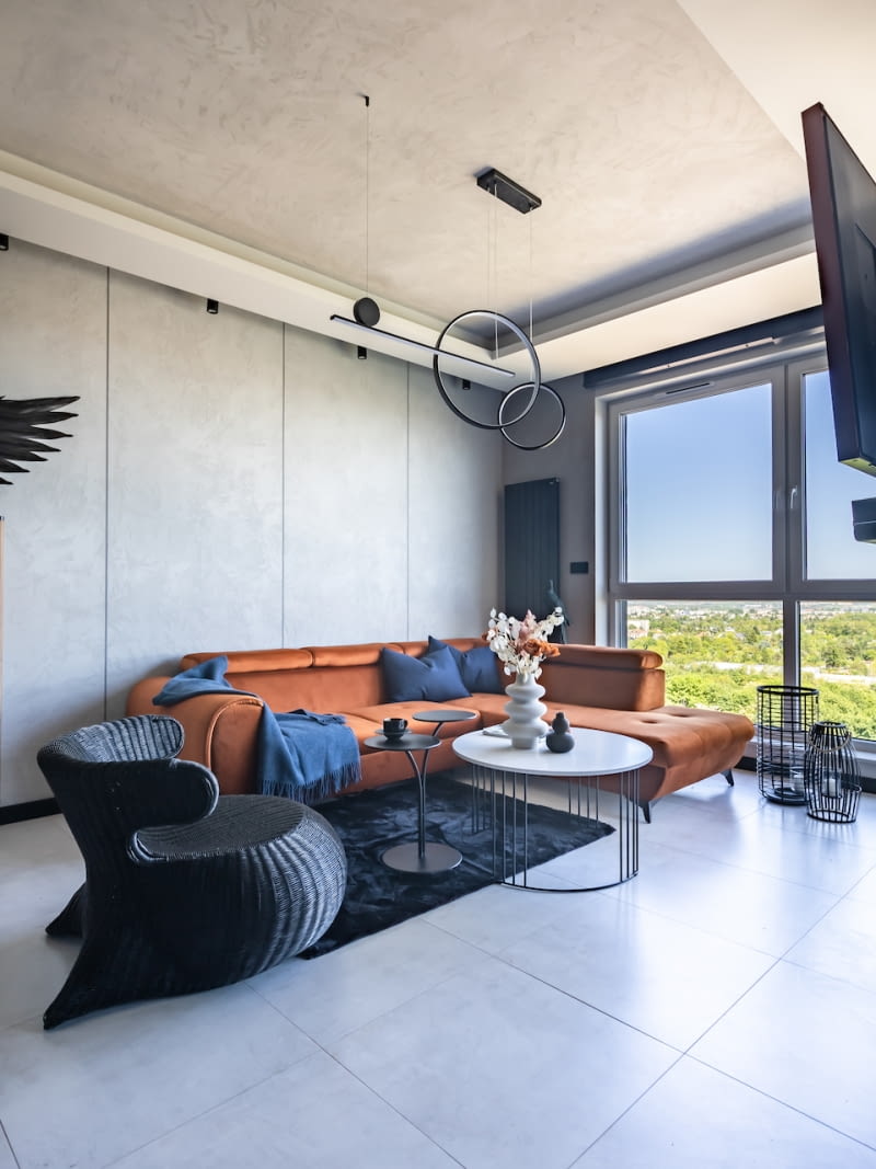 Wystrój wnętrz mieszkania podkreśla podział na dwie strefy funkcjonalne – dzienną i nocną. W pierwszej, reprezentacyjnej części dominują odcienie szarości od jasnych, po antracytowe. Szara podłoga z płytek ceramicznych i szare, pokryte ozdobnym tynkiem ściany oraz wnęki w sufitach podwieszanych pięknie eksponują dobrane do aranżacji rude i drewniane elementy. W części nocnej do przyjętej palety barw dołączają beże i odcienie niebieskiego. Nadają aranżacji łagodniejszego, niż w surowej części dziennej, klimatu.