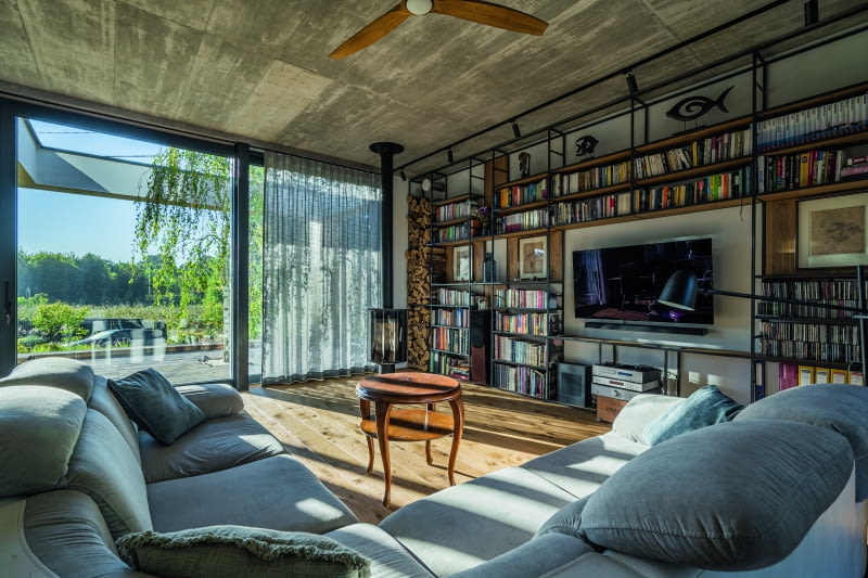 Połączone ze sobą salon, jadalnia i kuchnia pełne są akcentów przemysłowych, na które składają się zarówno betonowy sufit, uzyskany z szalunku  stropu, jak i stylowa żeliwna koza. Elementami ocieplającymi ów wizerunek są wygodne meble z miękkim, szarym obiciem i „antykwaryczny” stolik  kawowy, a gdzieś pośrodku tych klimatów lokuje się duma właścicieli – robiony na zamówienie regał na całą ścianę, wykonany z metalu i drewna