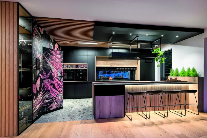 W otwartej na salon kuchni znowu drewniane lamelki – tym razem nie tylko położone na suficie, ale i obecne na ścianie. Do tego zadbano  o wyraziste akcenty kolorystyczne na powierzchni, w której wykrojono drzwi wiodące do niedużej spiżarni.